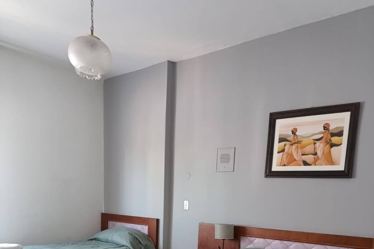 A hotel room with two single beds, a wooden floor, a white pendant light, a framed painting, and a bedside table with a lamp.