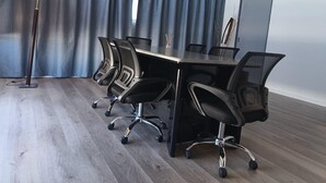 Un salón de conferencias con una mesa larga, cuatro sillas de oficina negras, una lámpara de pie y una cortina gris.