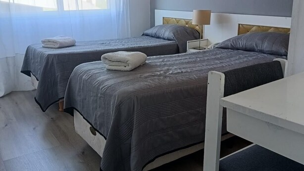 Habitación de hotel con dos camas, una mesa blanca y una ventana con cortinas translúcidas.