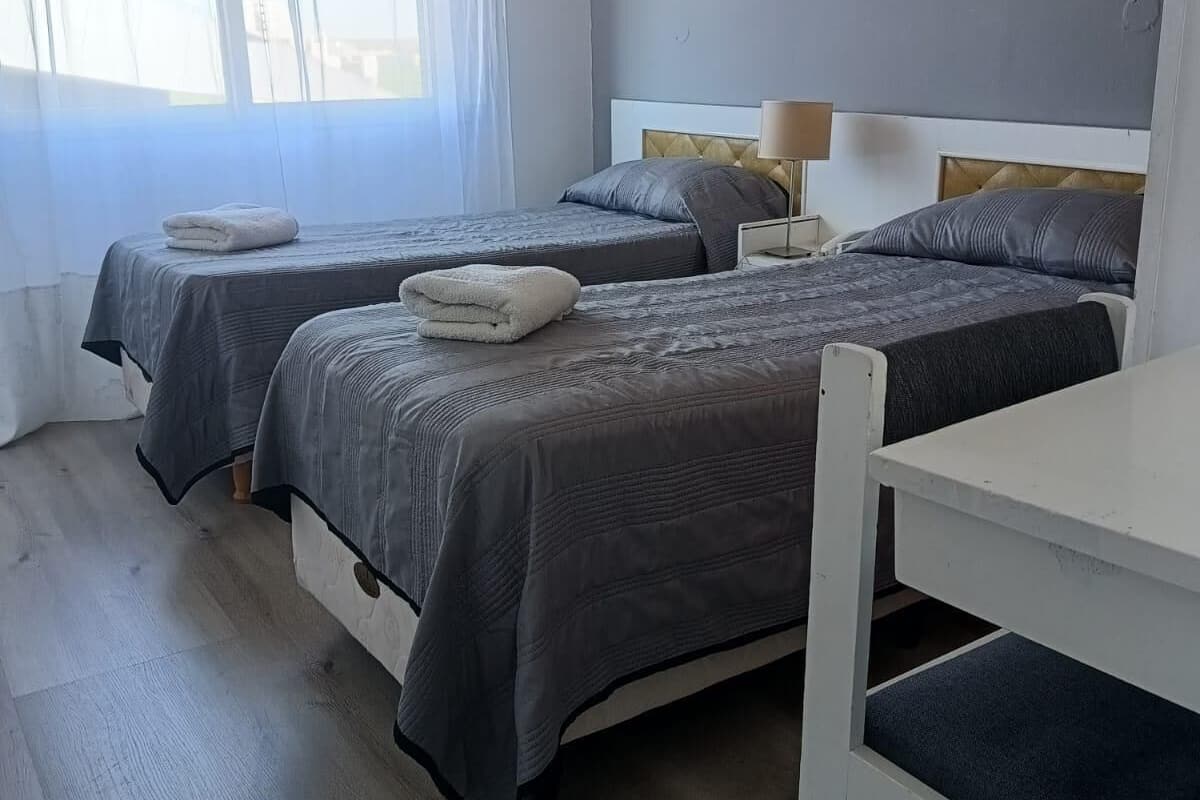 A hotel room with two beds, a white table, and a window with sheer curtains.