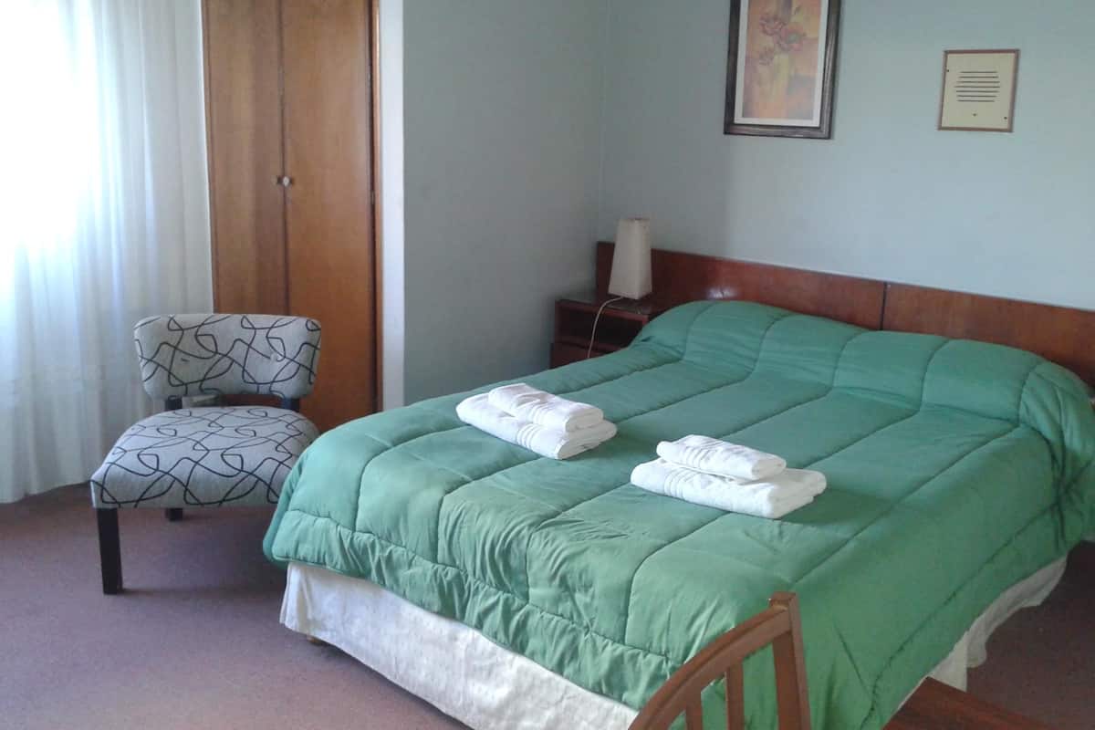 A neatly made bed with green bedding and two folded towels, a patterned armchair, a wooden door, and a framed picture on the wall.