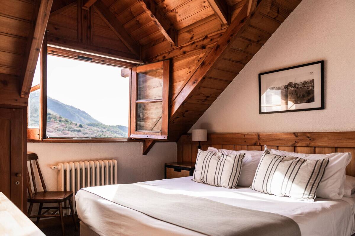Ein gemütliches Schlafzimmer mit Holzbalkendecke, einem großen Bett mit gestreiften Kissen, einer Heizung, einem Sessel und einem Fenster mit Bergblick.
