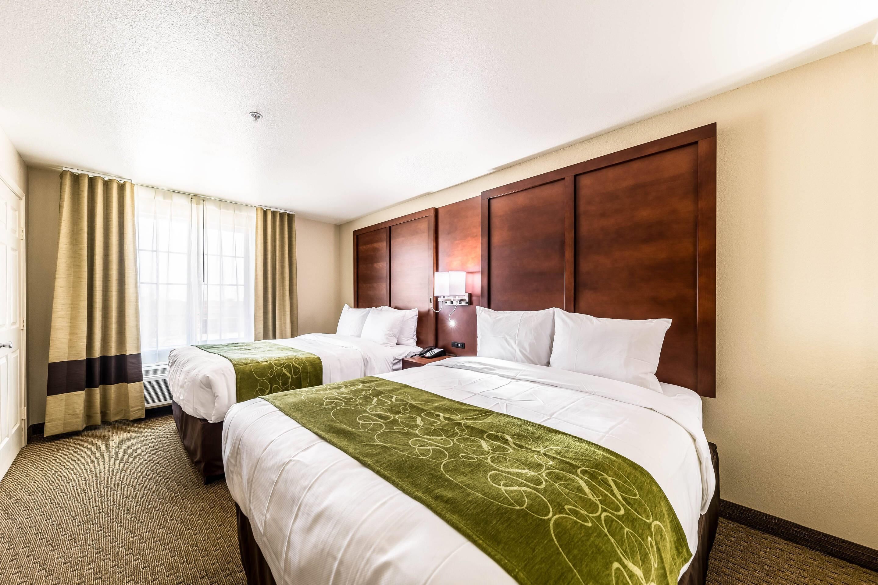 Ein Hotelzimmer mit zwei Betten, einem großen Holzkopfteil, einem Schreibtisch mit Lampe und einem Fenster mit Vorhängen.
