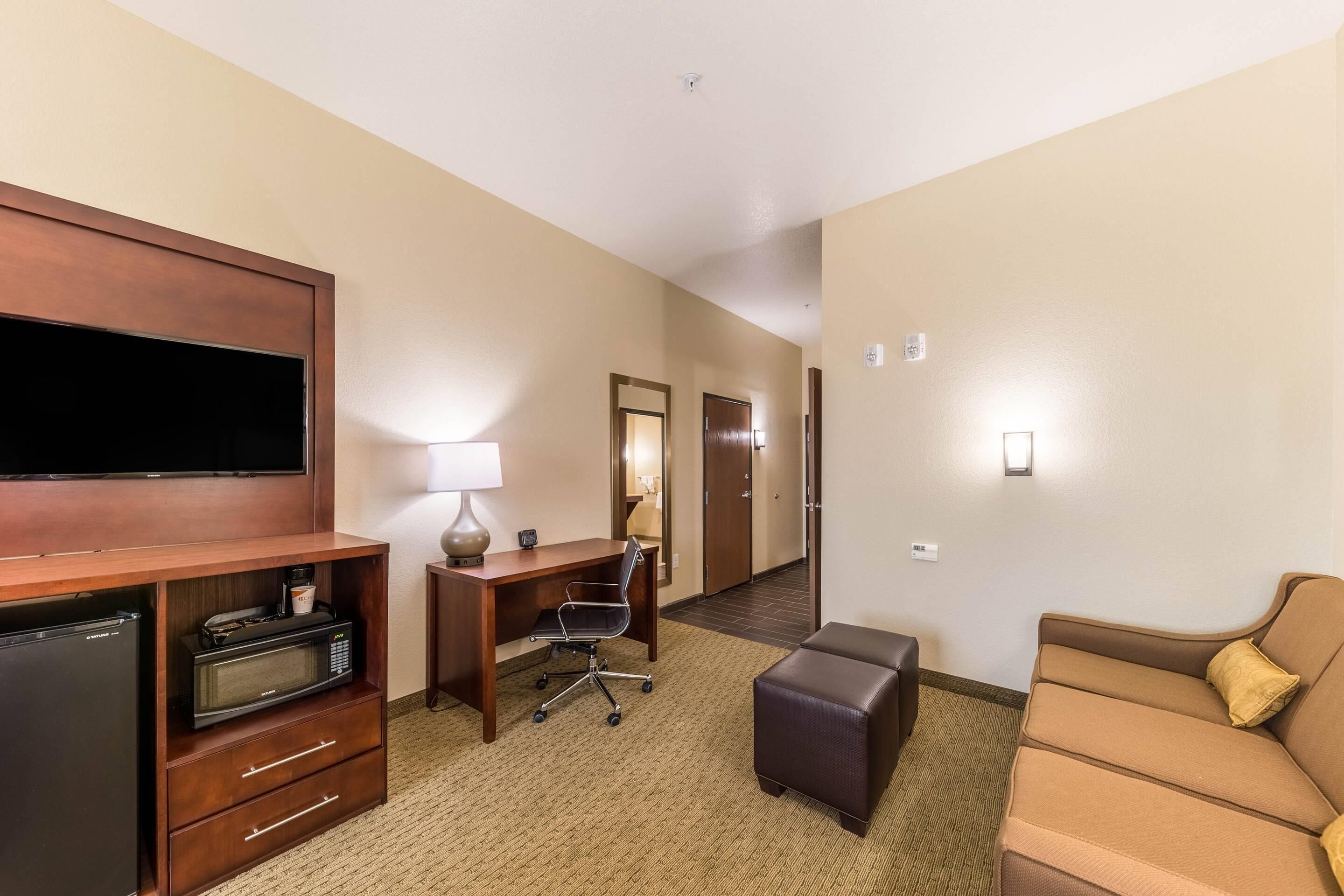 A hotel room with a flat-screen TV, a desk with a lamp, a microwave, a refrigerator, a brown leather sofa, and a hallway leading to a door.