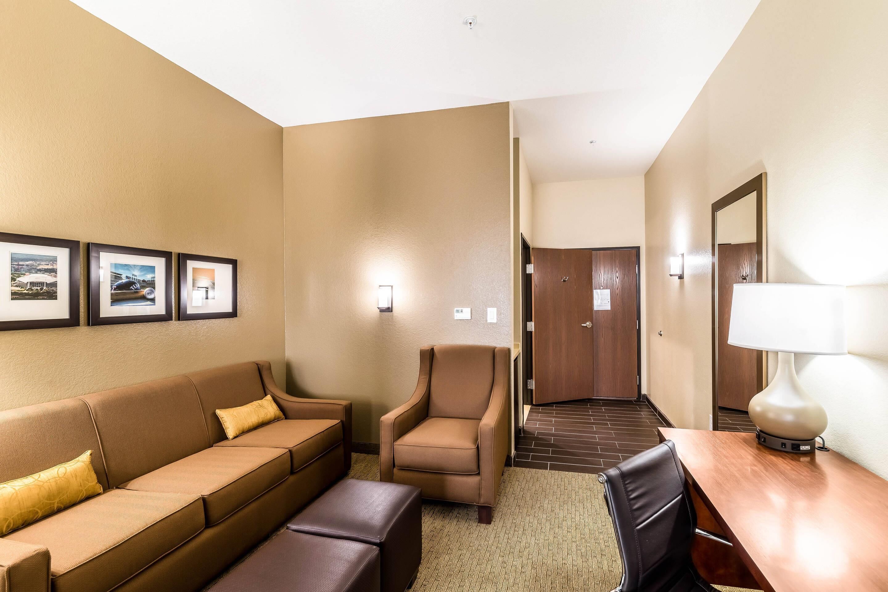 Une chambre d’hôtel avec un canapé marron, un fauteuil marron, une table en bois et une lampe. Des tableaux encadrés ornent le mur, et une porte donne accès à une autre pièce.