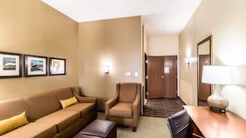 Habitación de hotel con un sofá marrón, un sillón marrón, una mesa de madera y una lámpara. Se observan cuadros enmarcados en la pared y una puerta que da a otra habitación.