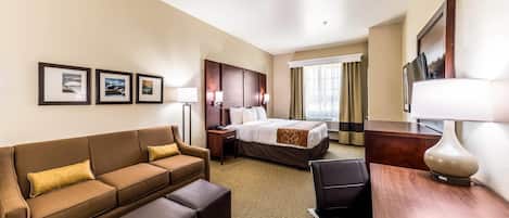 A hotel room with a brown sofa, a bed, a desk, and a chair. There are framed pictures on the wall and a window with curtains.
