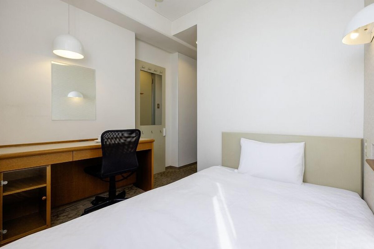 A hotel room with a bed, a desk, a chair, a mirror, and two wall-mounted lights.