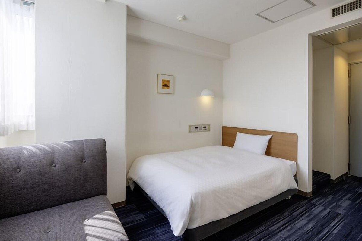 A hotel room with a bed, a grey sofa, a small bedside table, a wall-mounted light, and a window with sheer curtains.