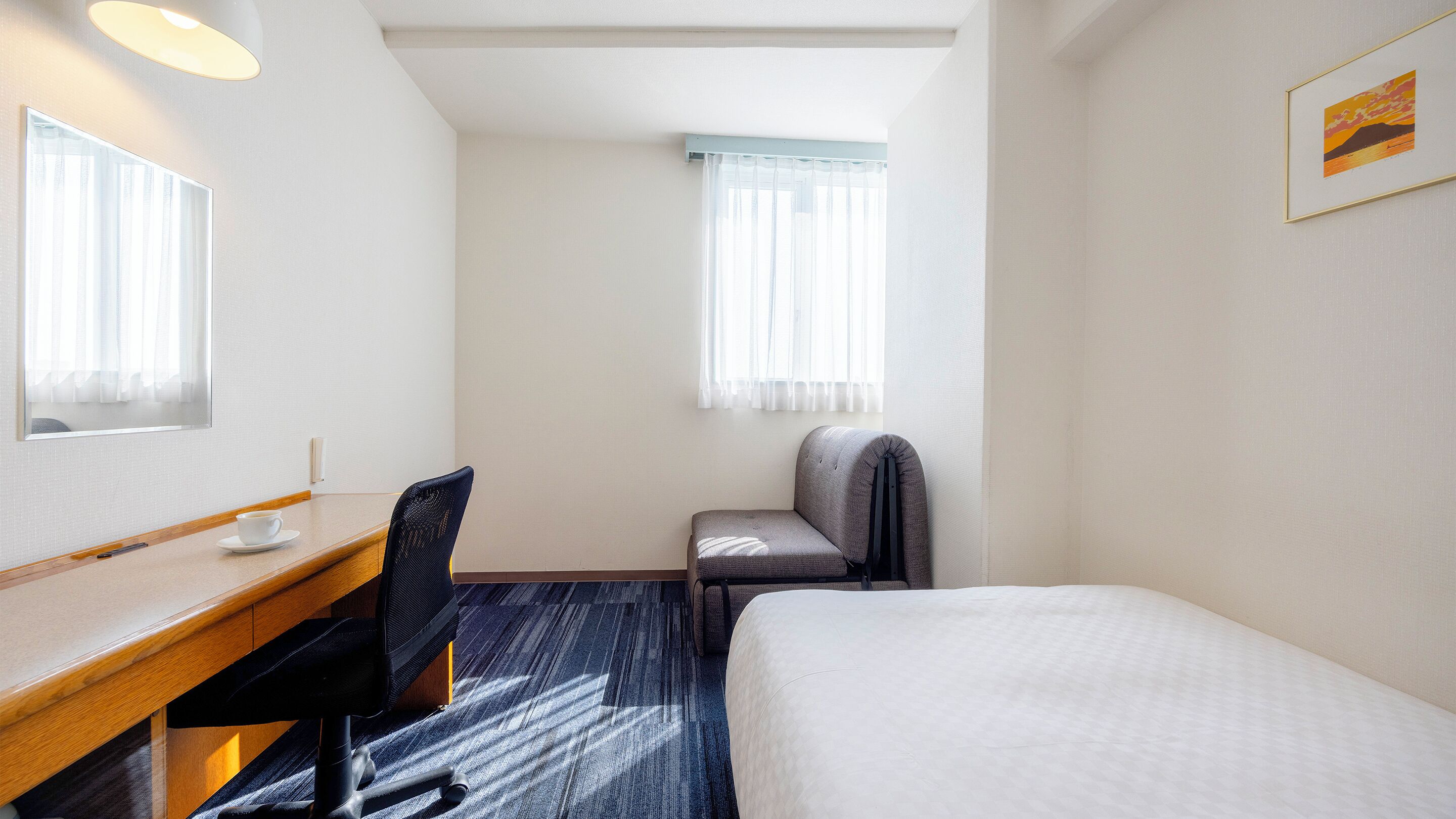 A hotel room with a bed, a desk, a chair, a window with curtains, and a framed picture on the wall.
