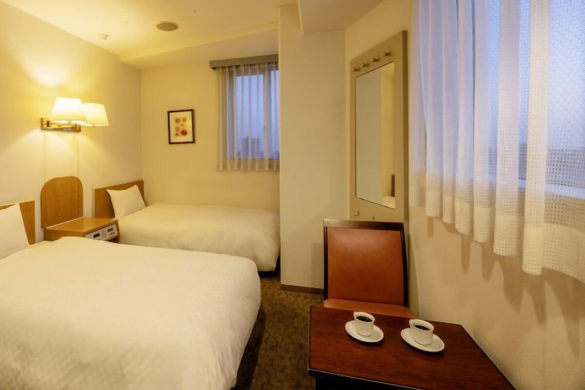 A hotel room with two beds, a bedside table with a lamp, a small table with two cups of coffee, a window with curtains, and a mirror on the wall.