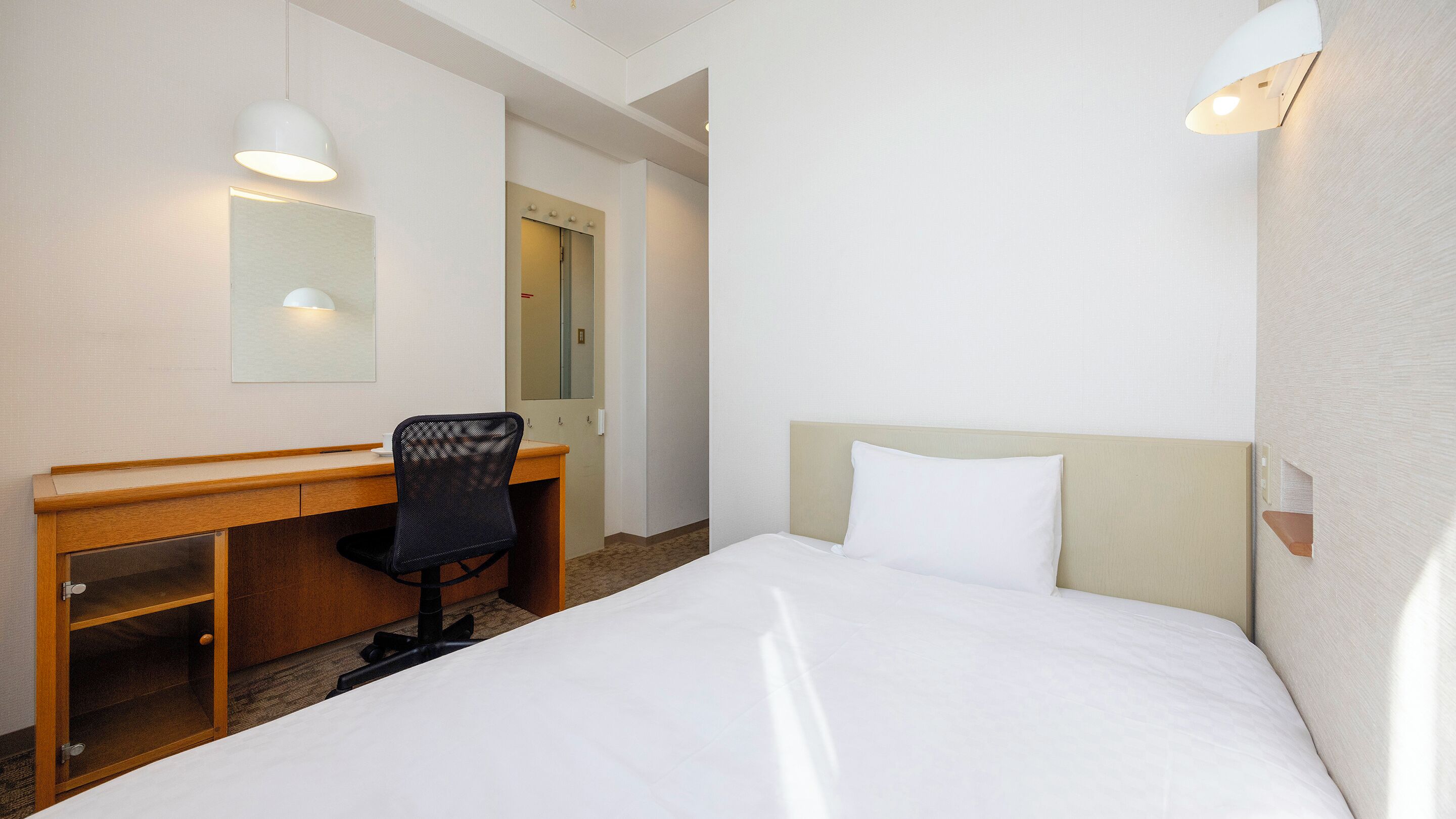 Habitación de hotel con cama, escritorio, silla, espejo y lámpara de pared.