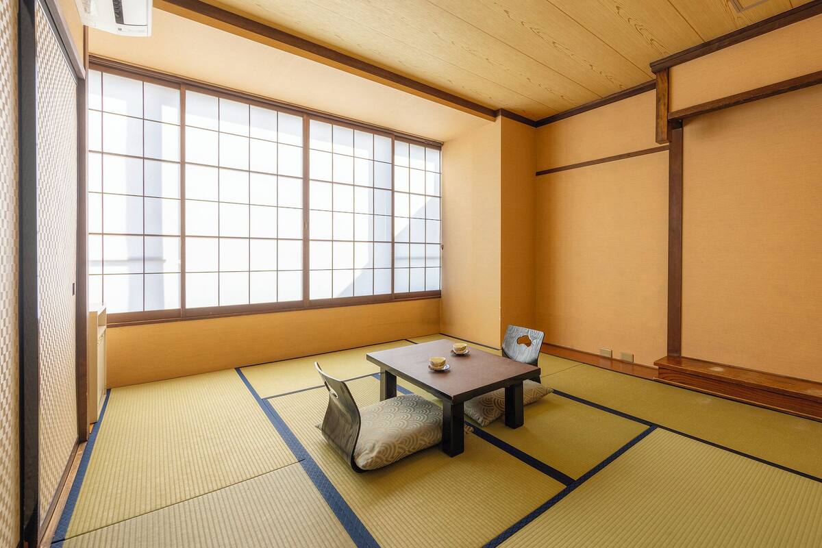 Une pièce avec un sol recouvert de tatamis, une petite table et des coussins, une porte coulissante et une grande fenêtre.