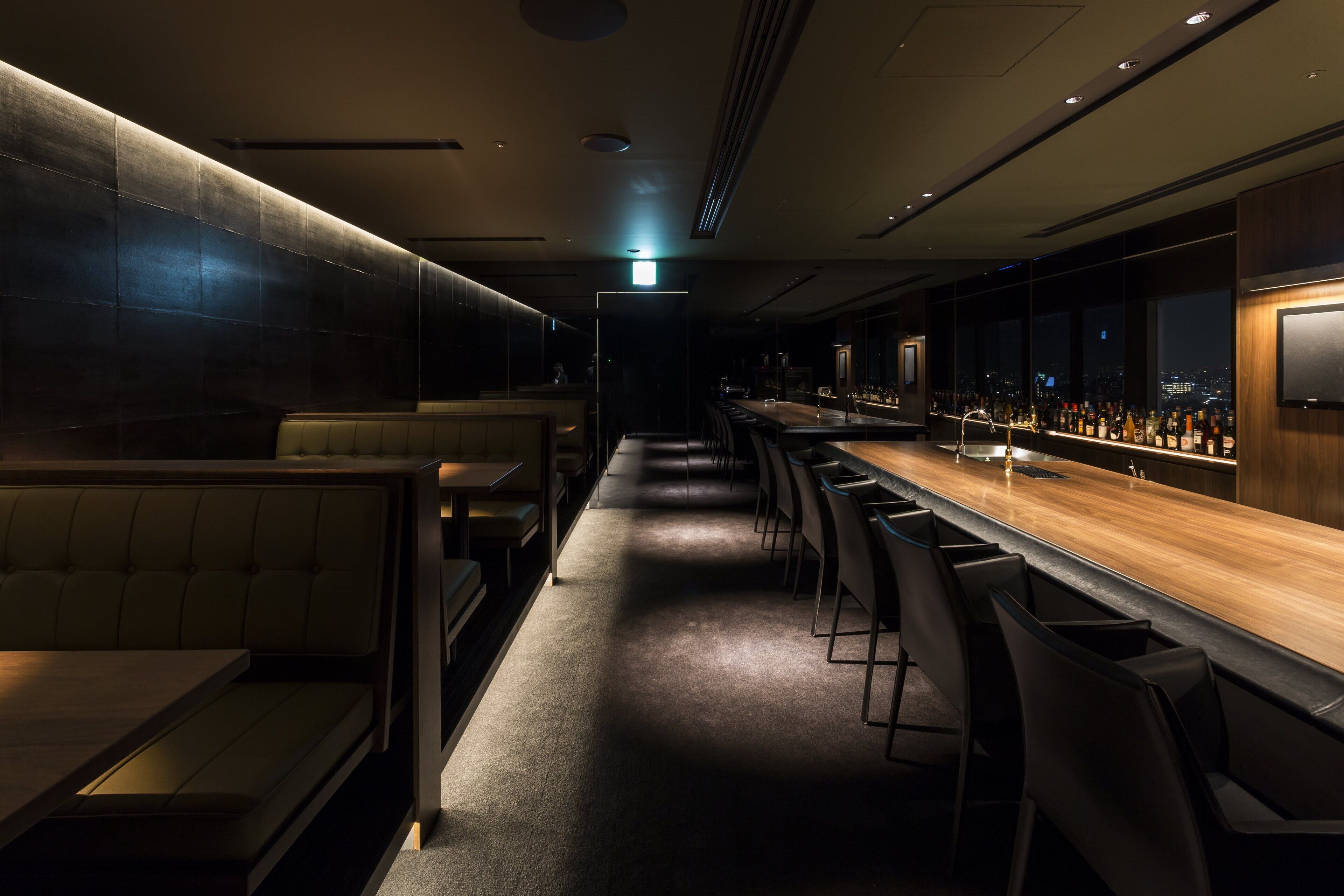 Uma área de bar com iluminação suave, um balcão comprido, banquetas e um espaço de estar com bancos estofados.