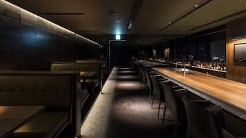 Uma área de bar com iluminação suave, um balcão comprido, banquetas e um espaço de estar com bancos estofados.