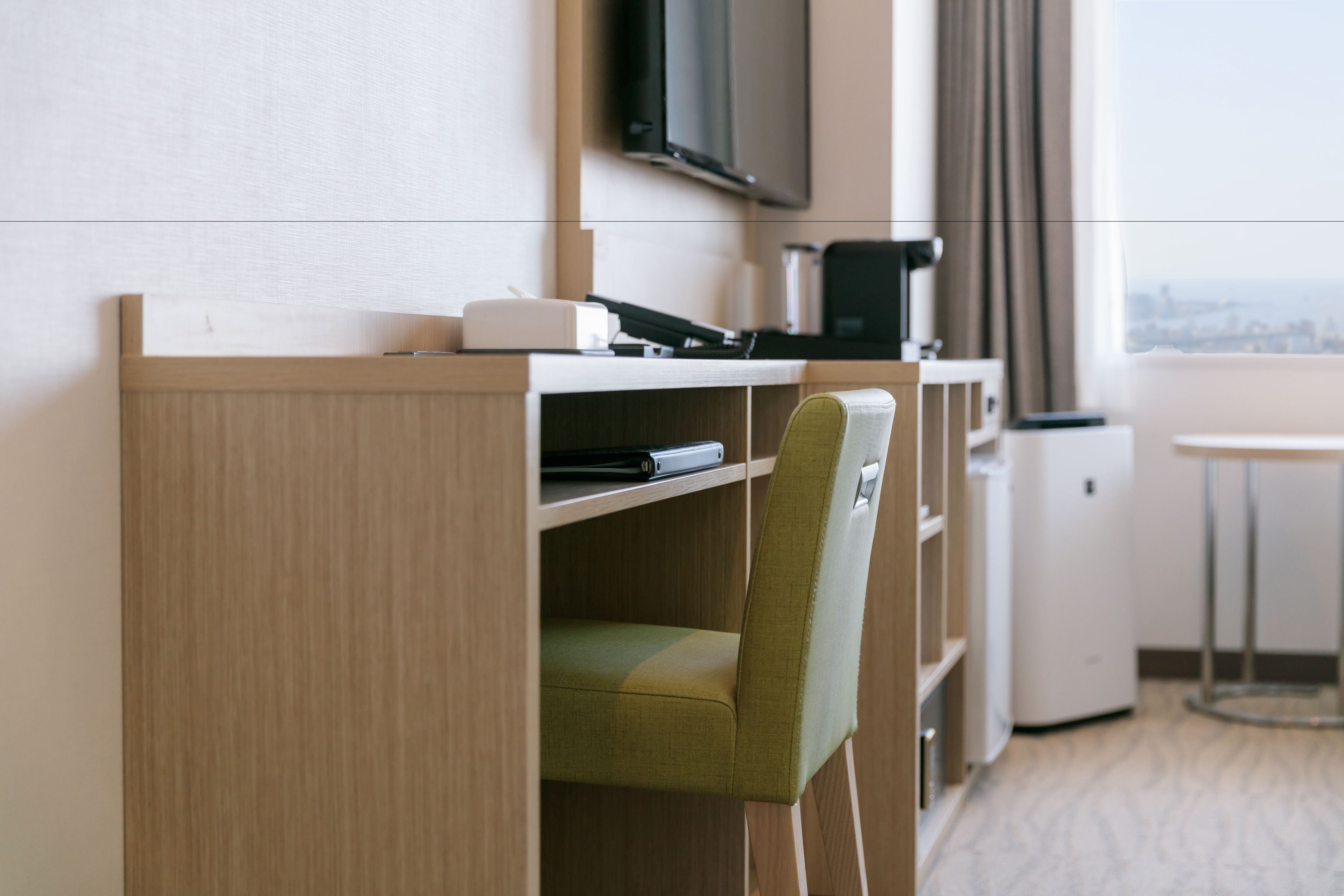 A hotel room with a wooden desk, a green chair, a flat-screen TV, a coffee machine, and a view of the city through the window.