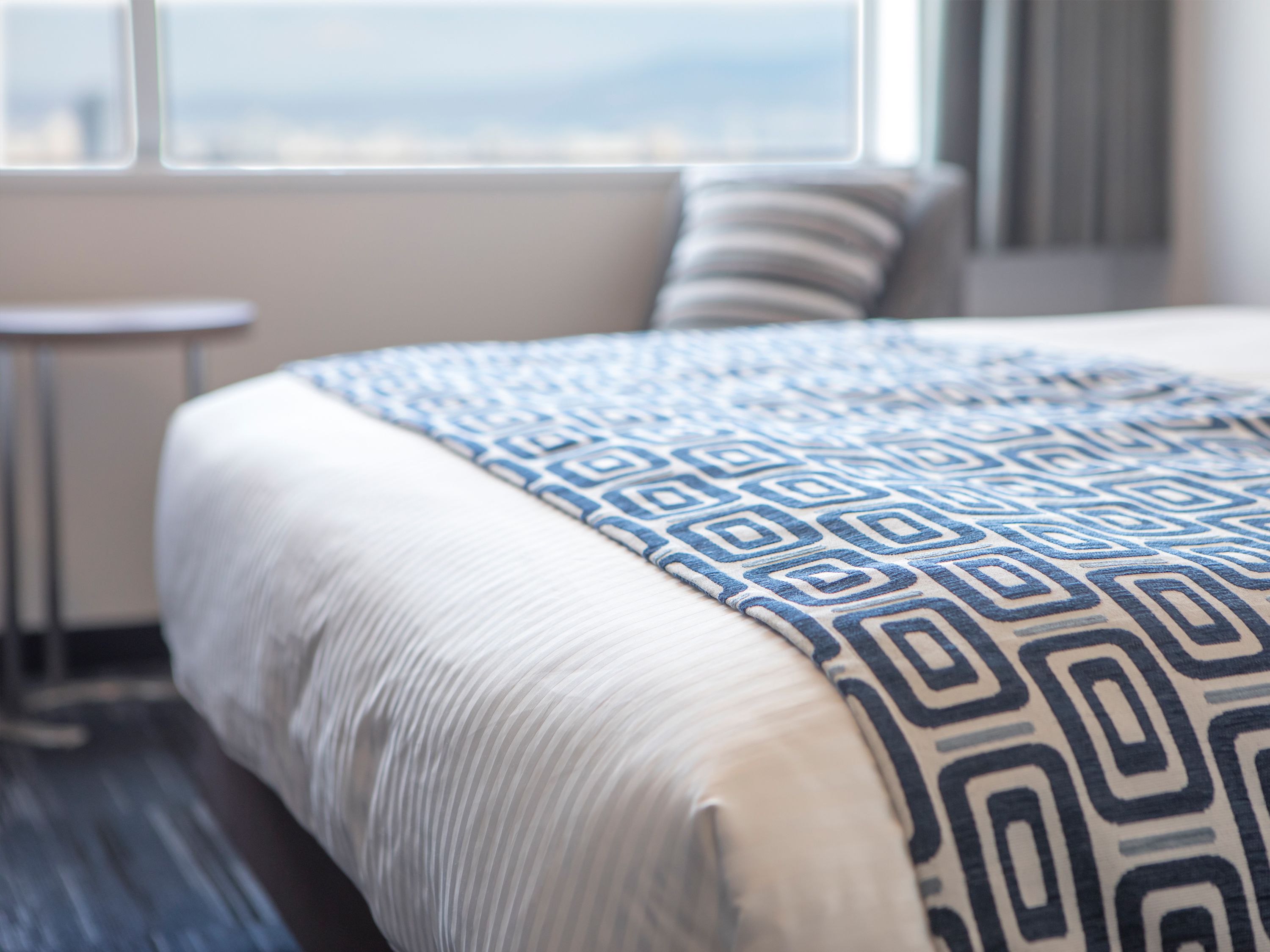 Een net opgemaakt bed met een bedsprei met patroon, een klein nachtkastje en een raam met uitzicht.