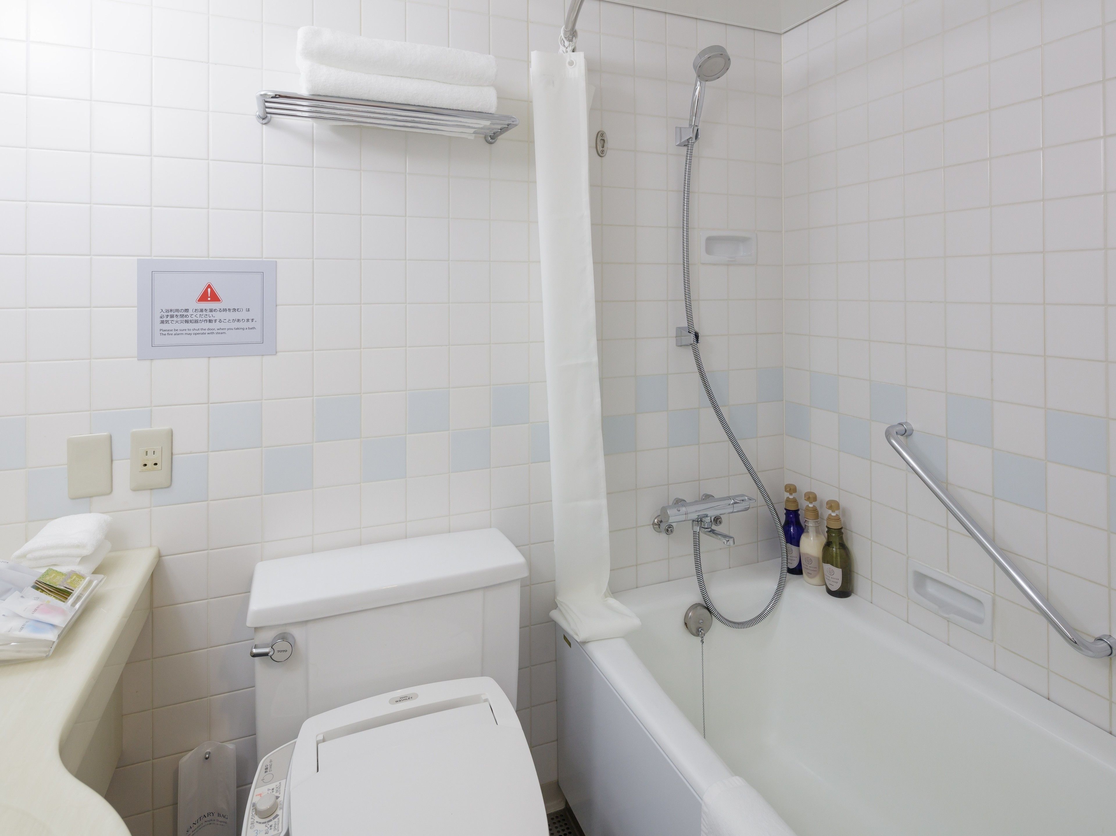 A bathroom with a toilet, sink, and bathtub. There is a showerhead and a grab bar. A sign with a warning symbol is visible on the wall.