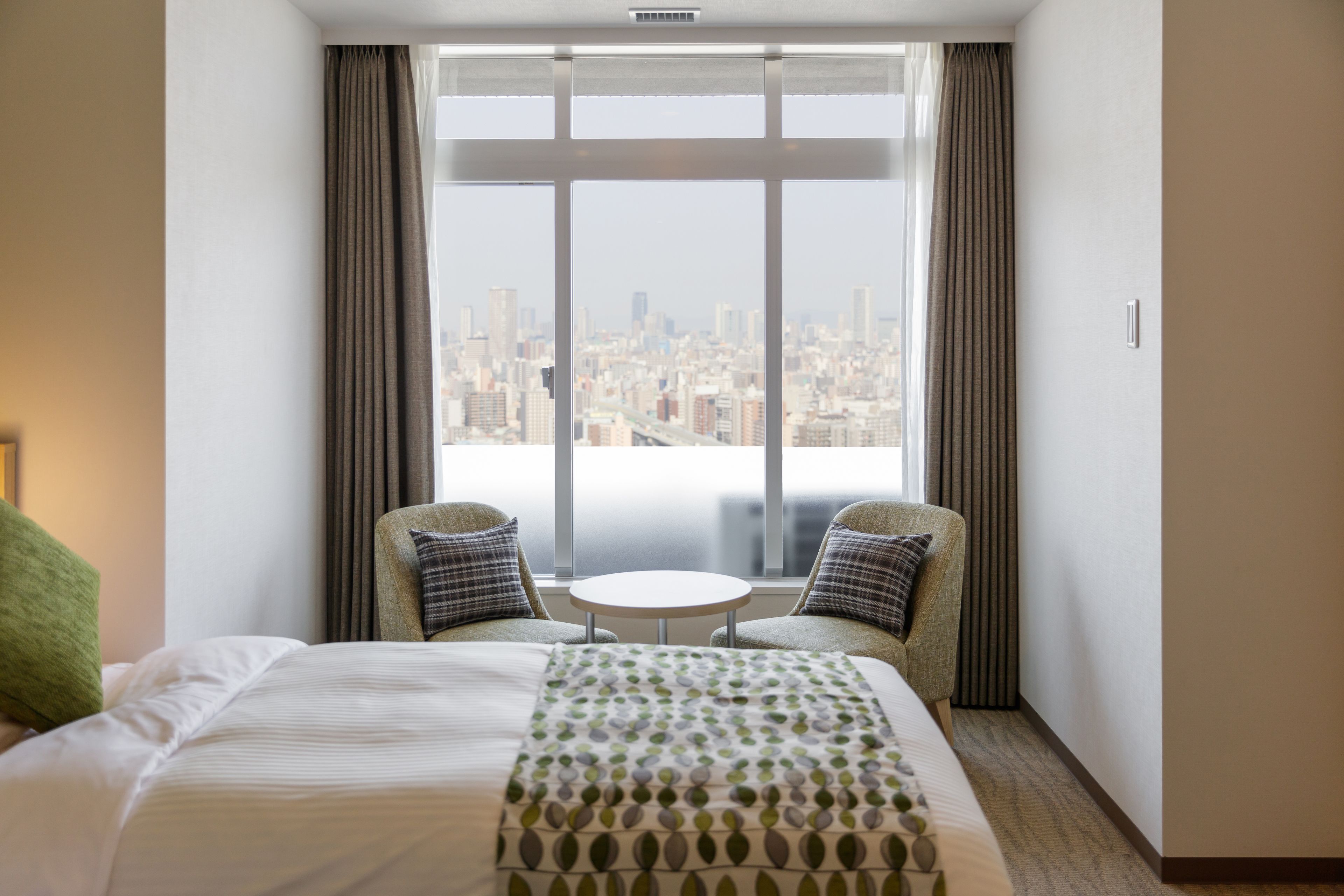 A hotel room with a large bed, two armchairs, a small table, and a city view through floor-to-ceiling windows.