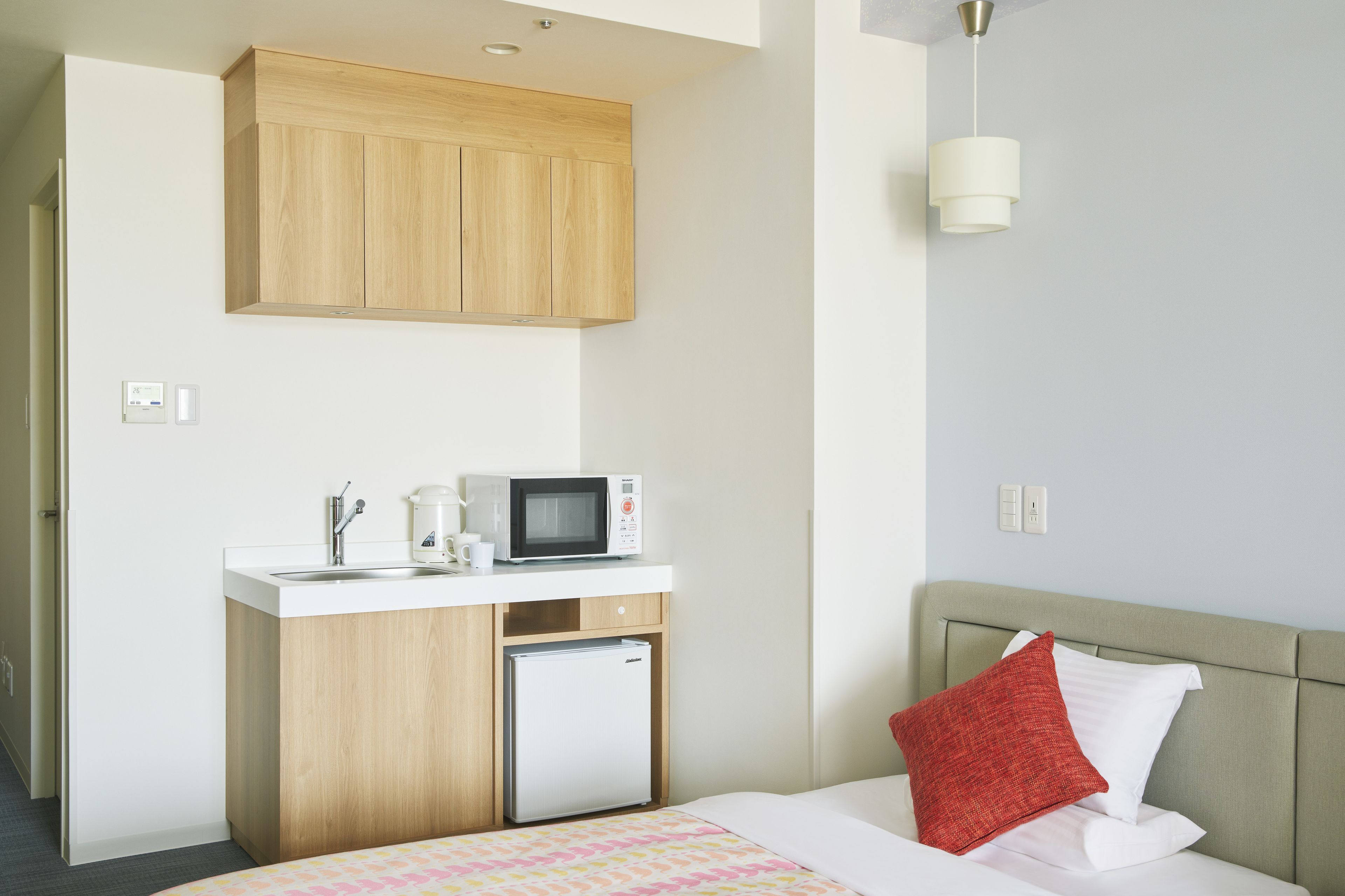 A compact hotel room with a kitchenette, a bed with a red pillow, and a light-colored headboard.