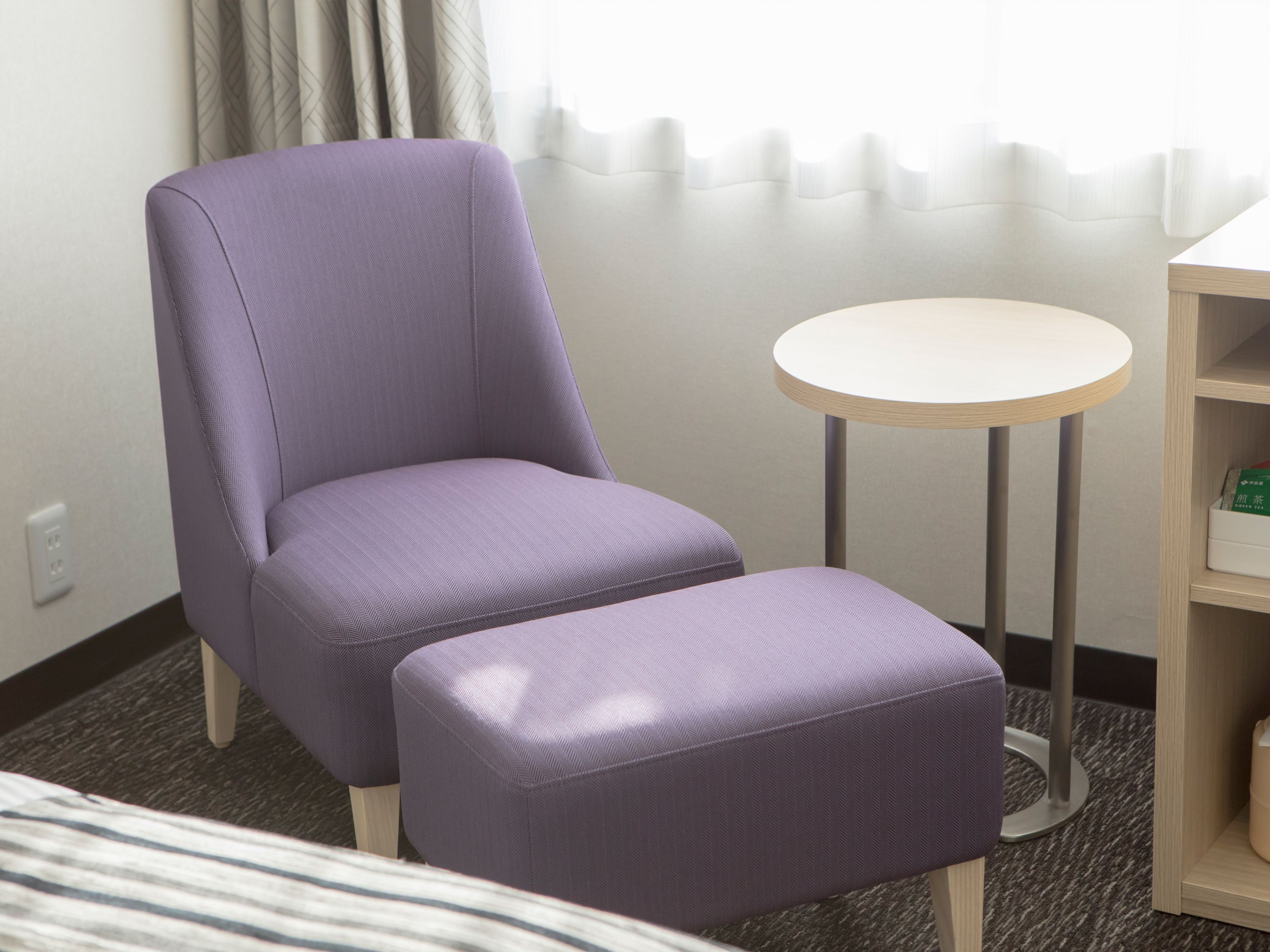 A room with a purple upholstered chair, a small round table, and a wooden bookshelf.
