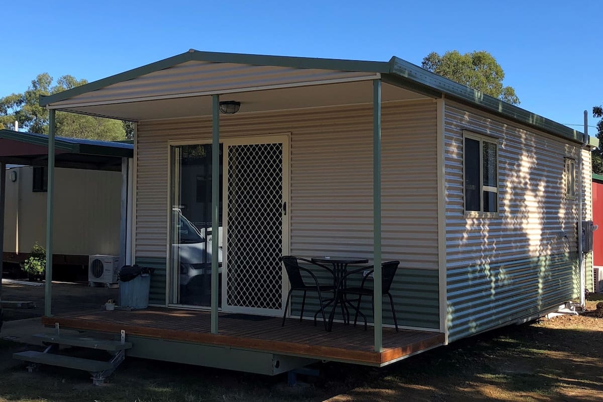 A small, single-story mobile home with a covered porch, a sliding door, and a small patio area with a table and chairs.