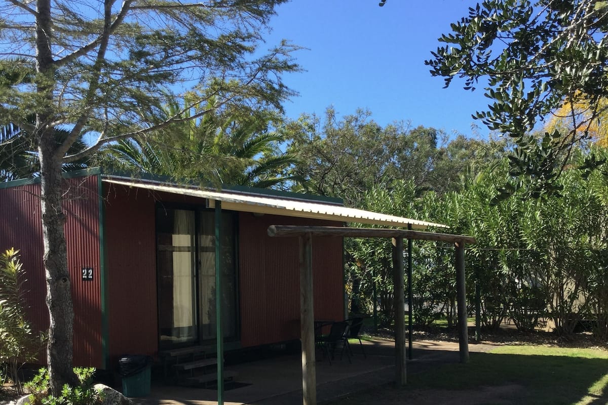 A small, single-story building with a metal roof and a green door, surrounded by trees and grass.