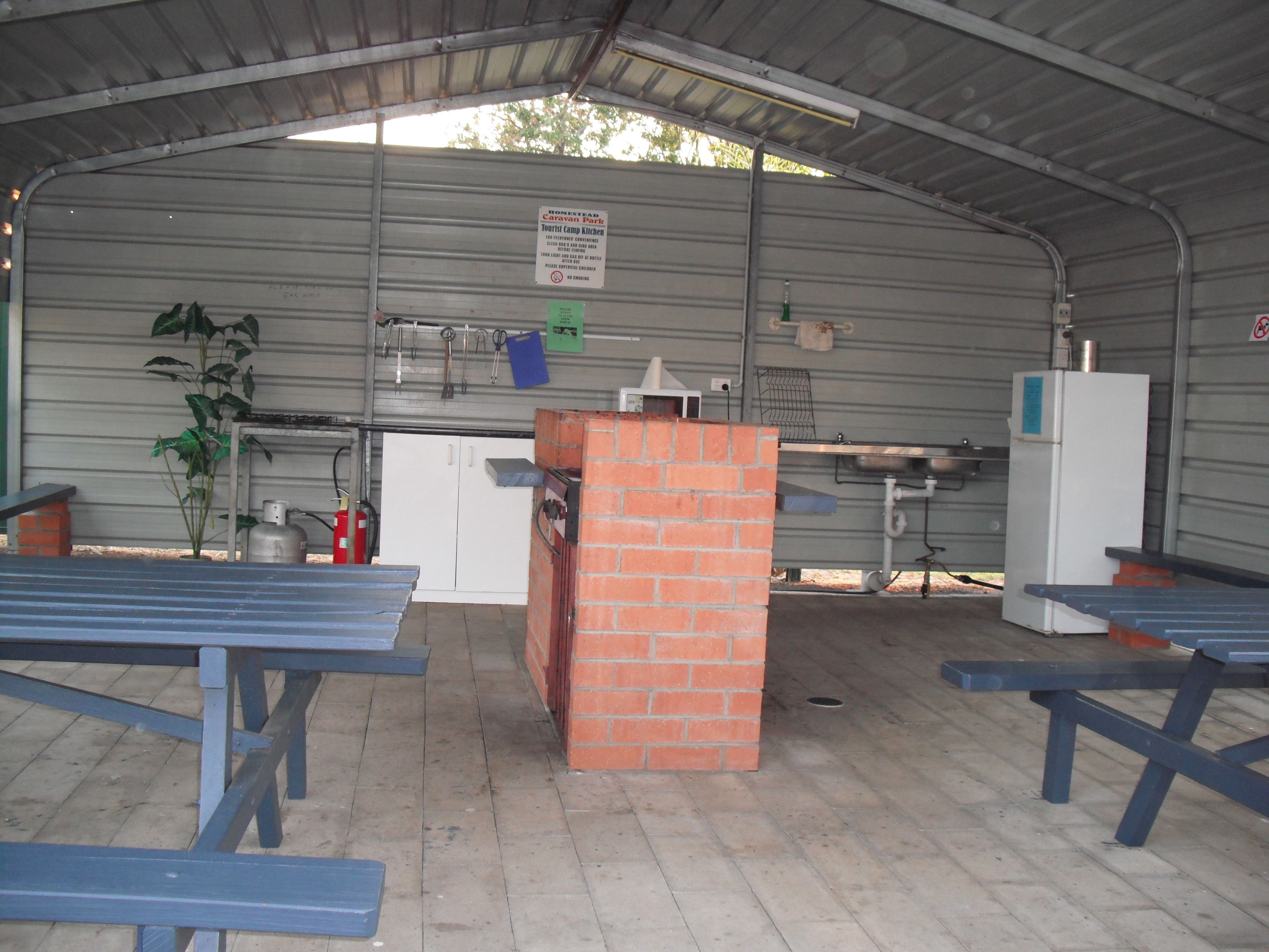 A covered outdoor area with picnic tables, a brick structure, and a kitchen area with a refrigerator.