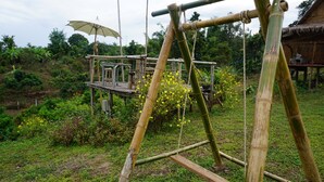 Ein Spielplatz mit einer Schaukel aus Bambus in einem üppigen Garten, im Hintergrund ein Gebäude mit einem Reetdach.