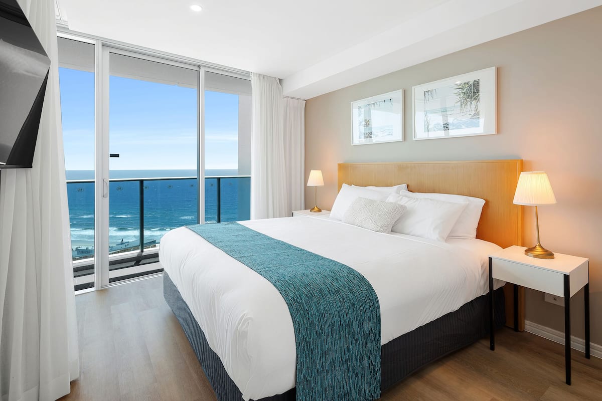 A hotel room with a large bed, a nightstand with a lamp, and a view of the ocean through a sliding door.