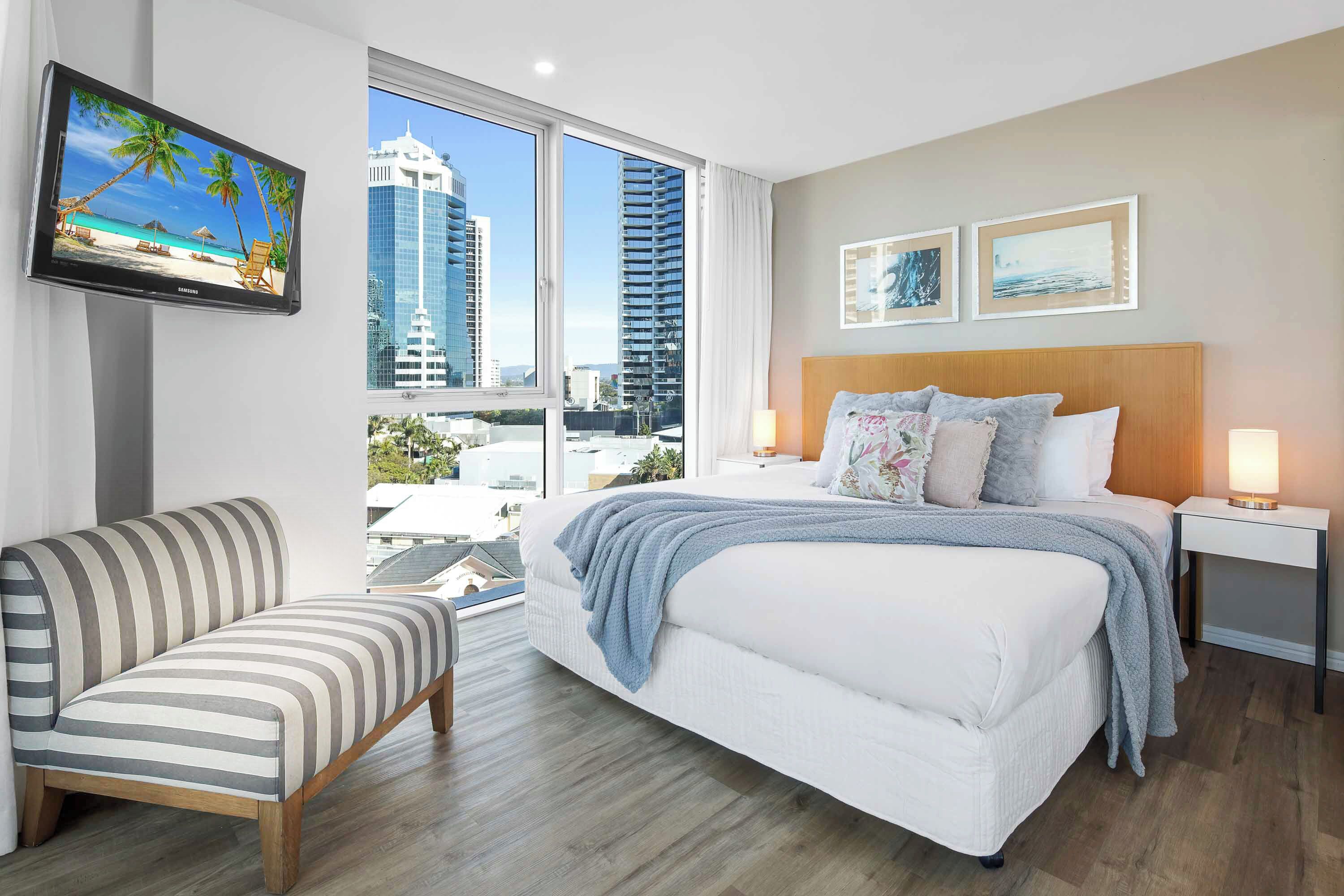 A hotel room with a bed, a TV on the wall, a window with a city view, and a chair.