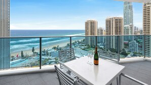 A balcony with a view of the beach and cityscape, a table with a bottle and glasses, and a railing.