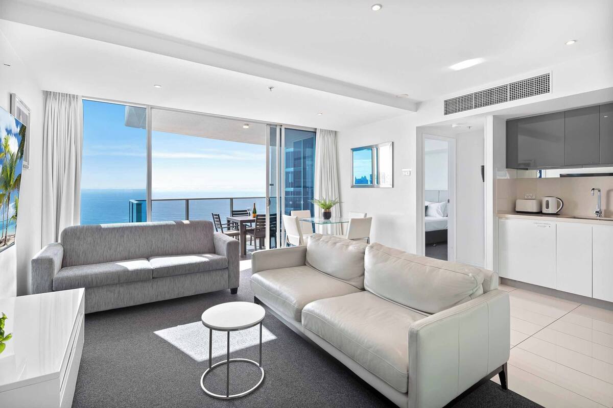 A modern living room with a grey sofa, a white sofa, a small round table, and a kitchen area with white cabinets and a flat-screen TV.