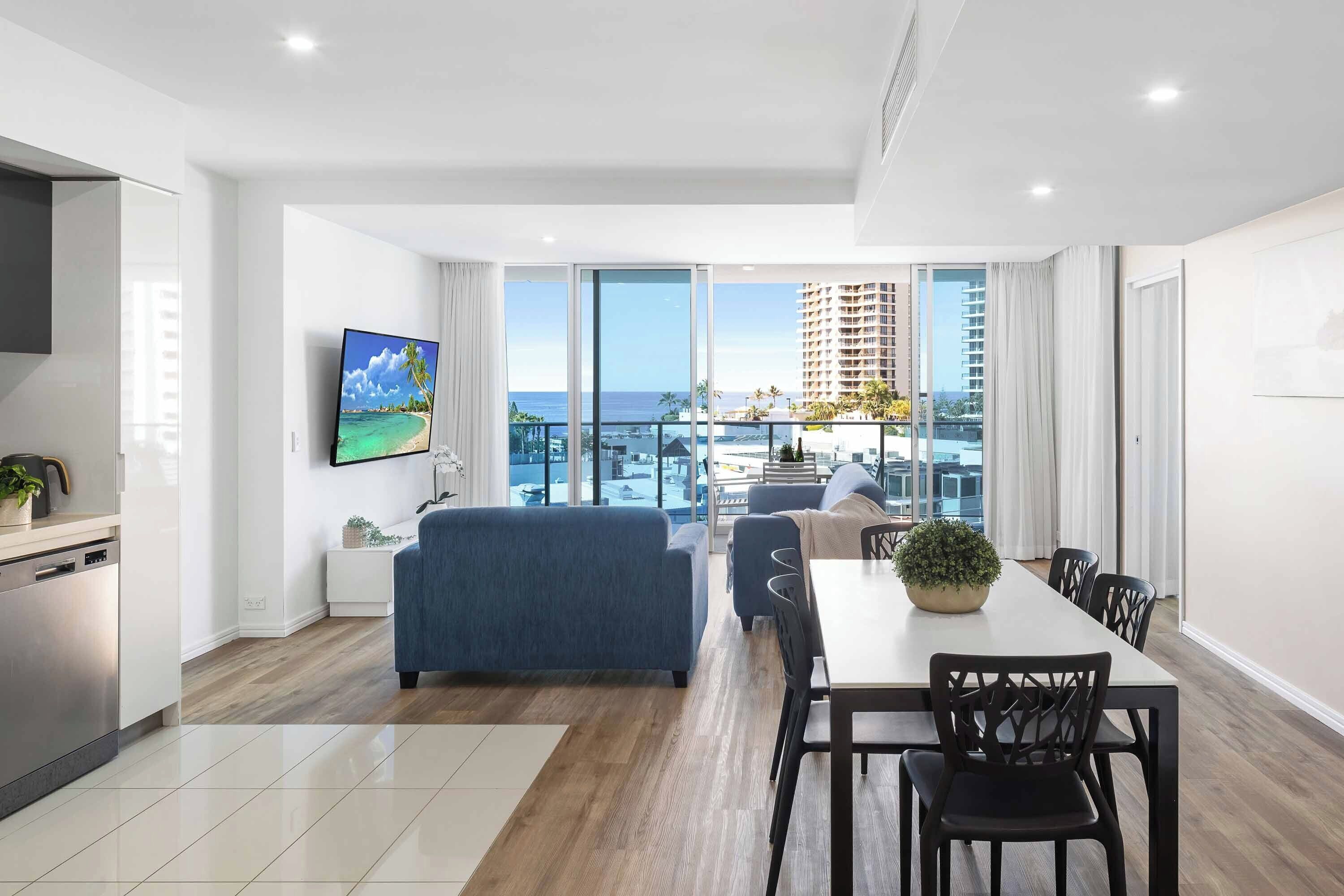 A modern living room with a large TV, a dining area with a table and chairs, and a balcony with a view of the city and ocean.