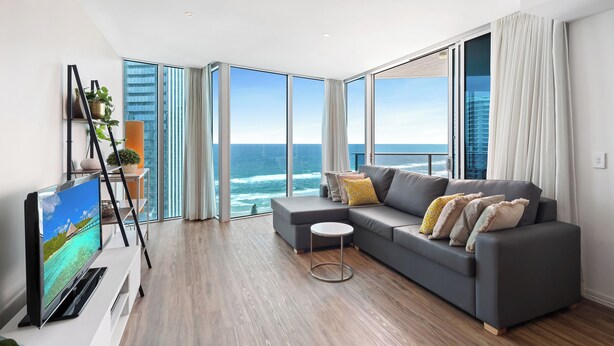 A modern living room with a grey sofa, a flat-screen TV, and a view of the ocean through large windows.