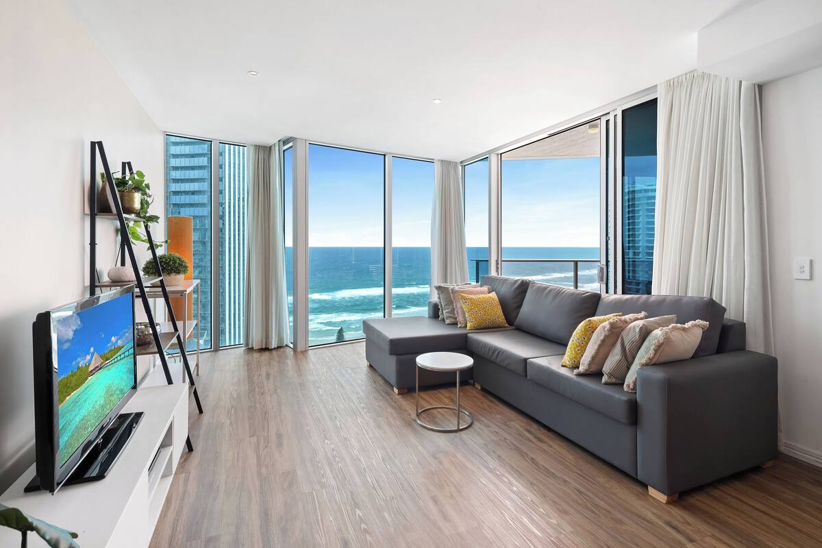 A modern living room with a grey sofa, a flat-screen TV, and a view of the ocean through large windows.