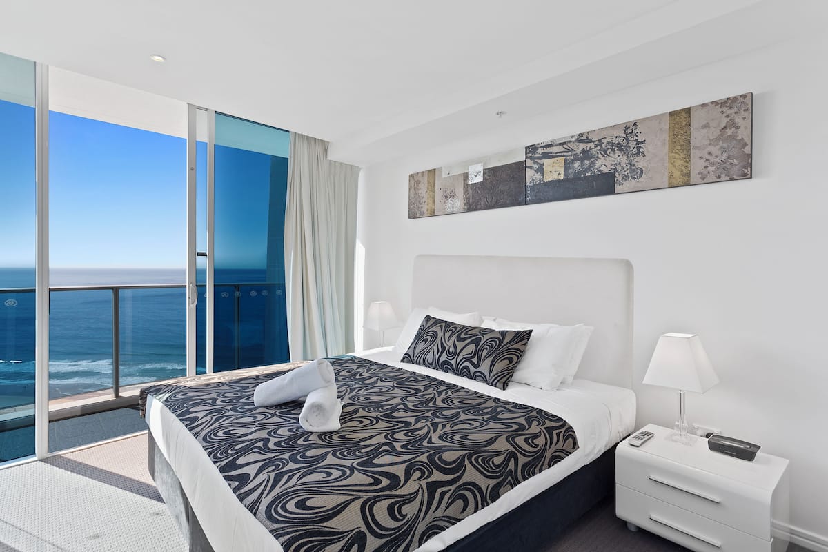 A hotel room with a large bed, a nightstand, a lamp, and a view of the ocean.