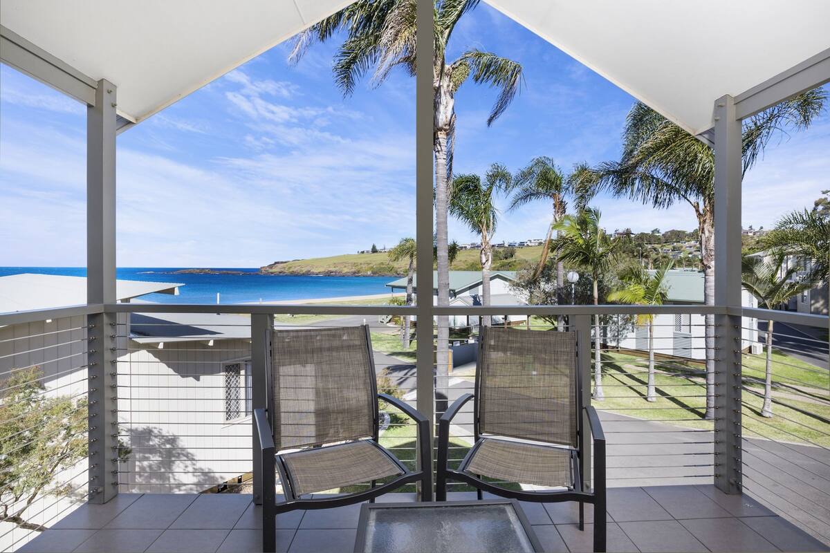A balcony with two chairs and a table, overlooking a beach and ocean.