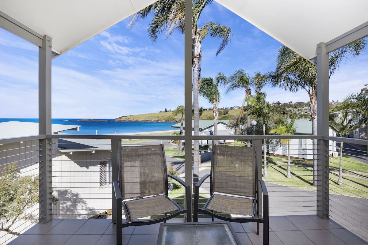 A balcony with two chairs and a table, overlooking a beach and ocean.