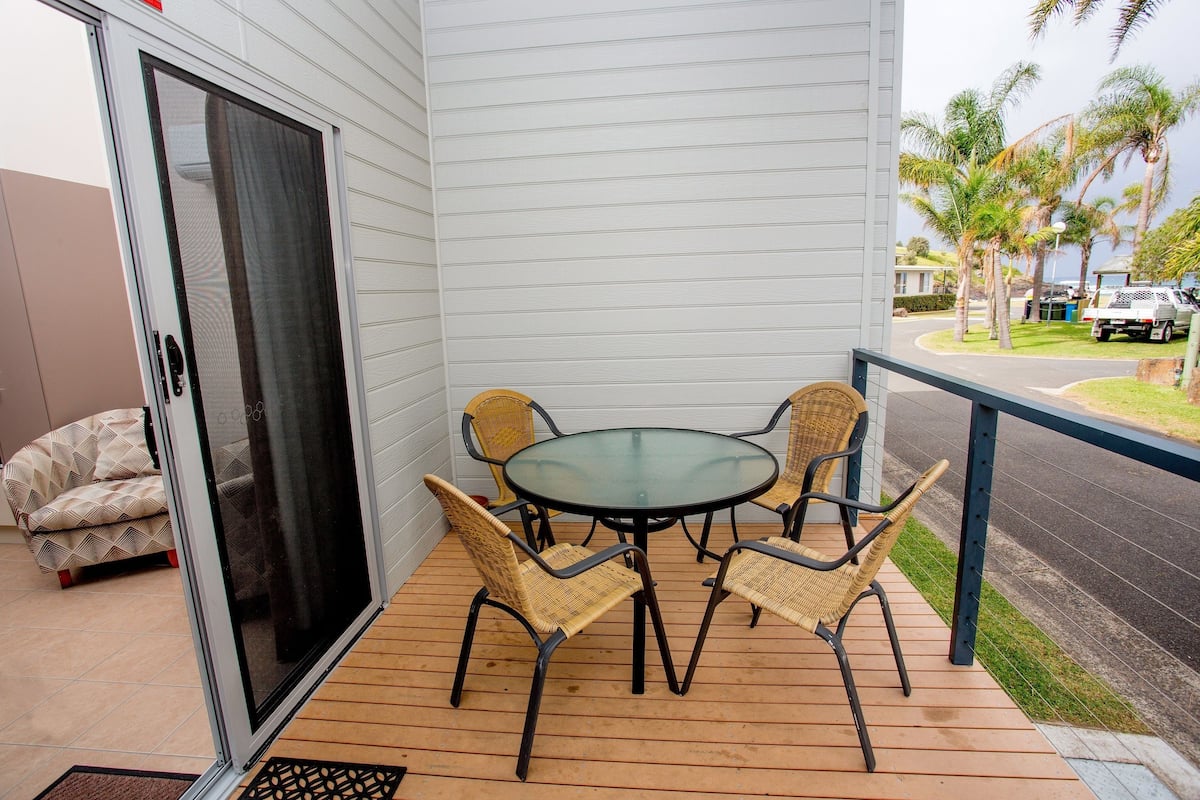 A covered patio with a glass table and wicker chairs, a sliding glass door, and a view of palm trees and a street.