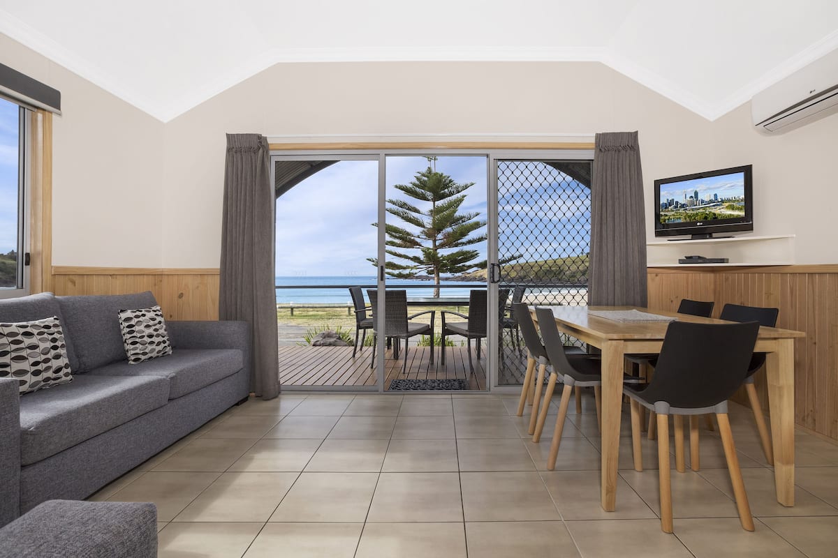 A modern living room with a grey sofa, a dining table with chairs, a TV mounted on the wall, and a large sliding glass door leading to a balcony with a view of the sea.