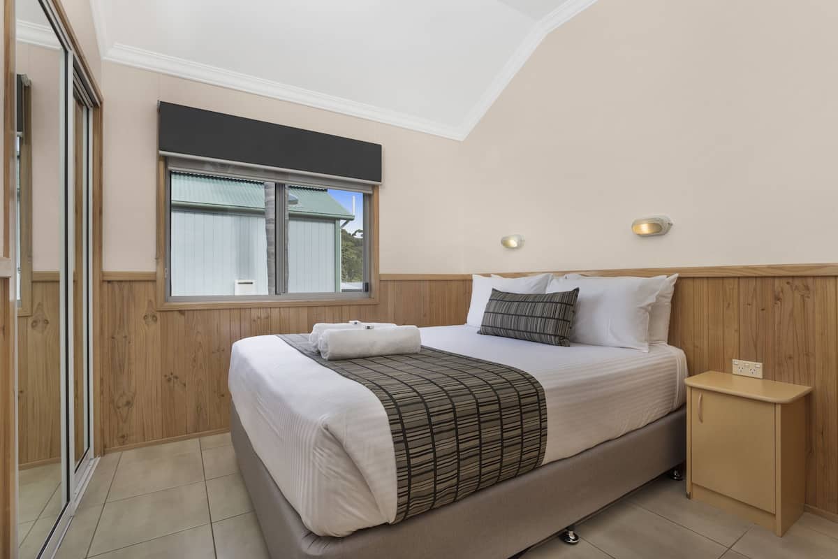 A bedroom with a bed, a nightstand, a window, and a wall-mounted light.