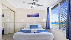 Quarto de hotel com uma cama, um ventilador de teto, uma janela com cortinas, um quadro na parede e vista para o mar.