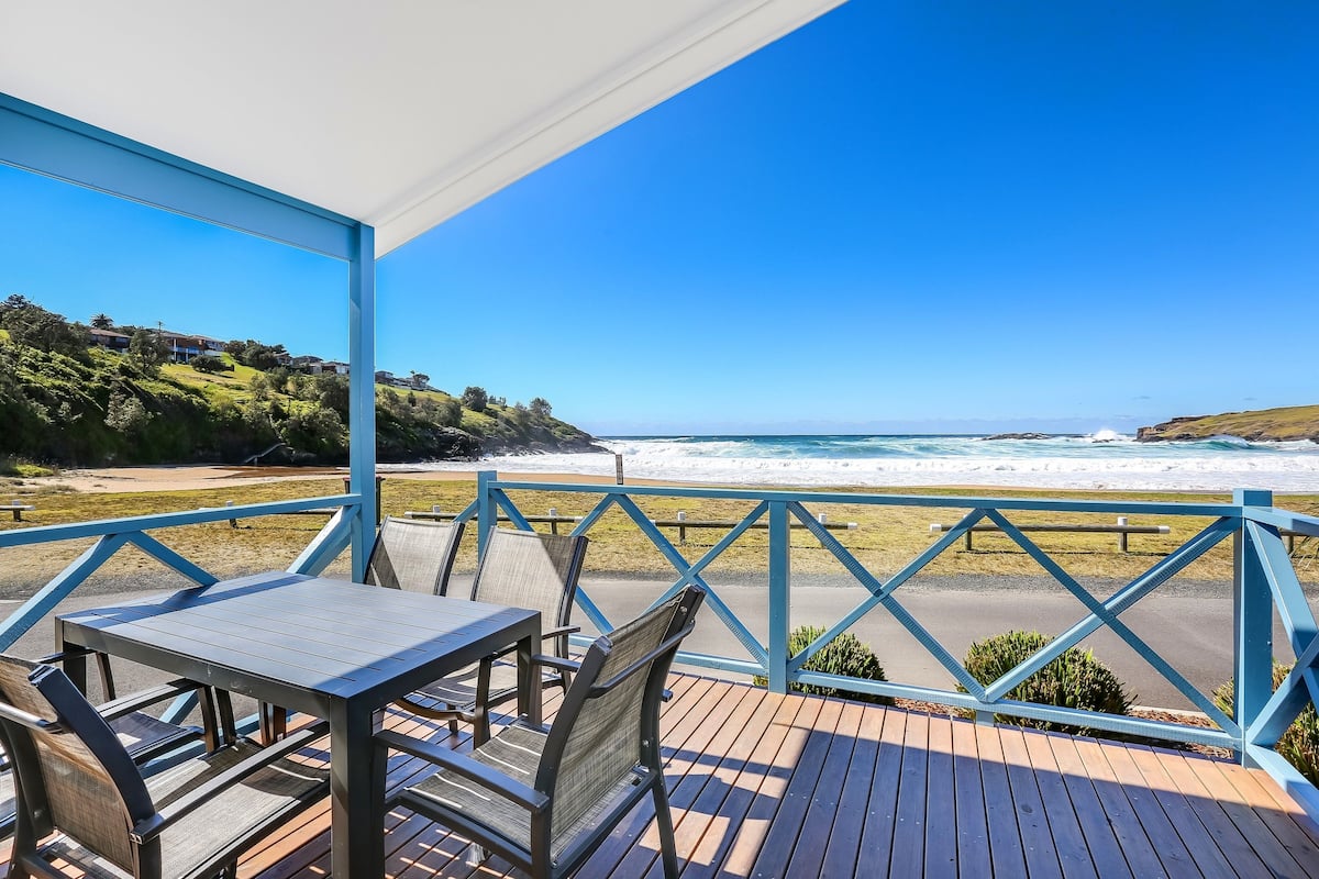 A wooden deck with a table and chairs overlooking a beach with waves.