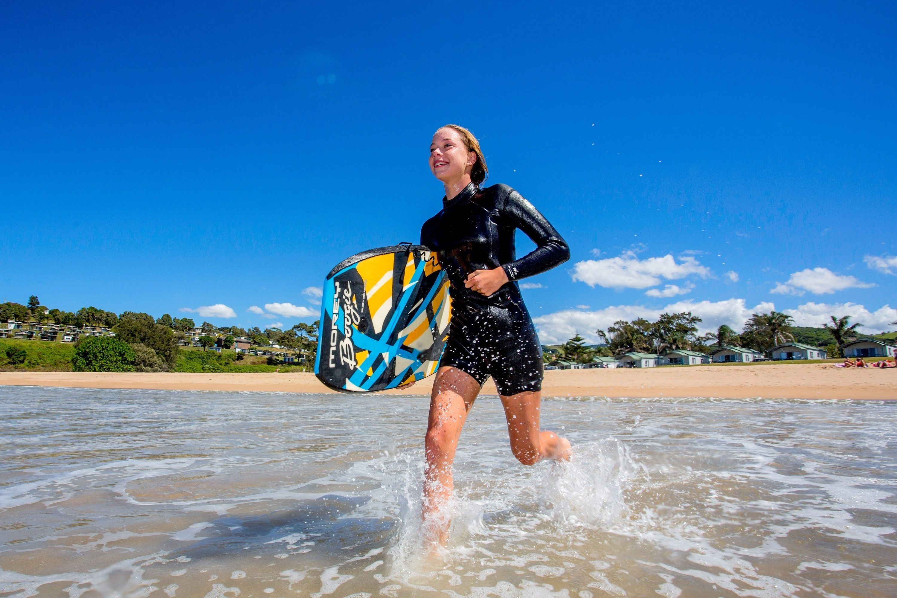A person in a wetsuit carrying a surfboard, walking through shallow water at a beach with houses in the background.