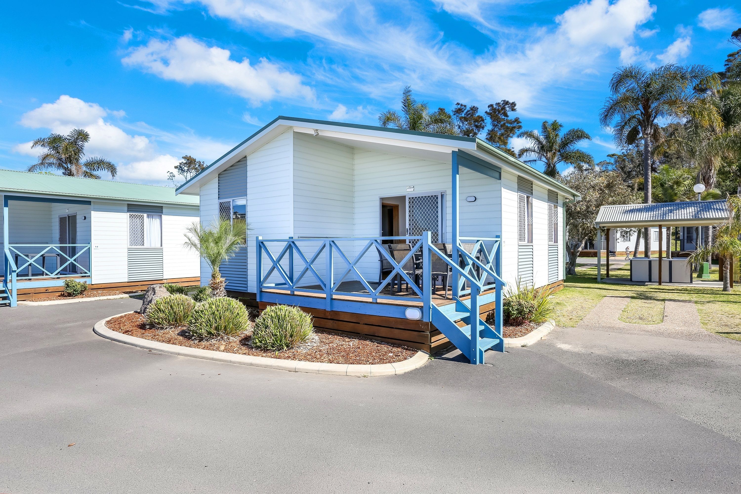 A single-story mobile home with a porch and a small deck, surrounded by a landscaped garden and palm trees.