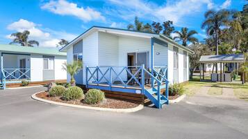 A single-story mobile home with a porch and a small deck, surrounded by a landscaped garden and palm trees.