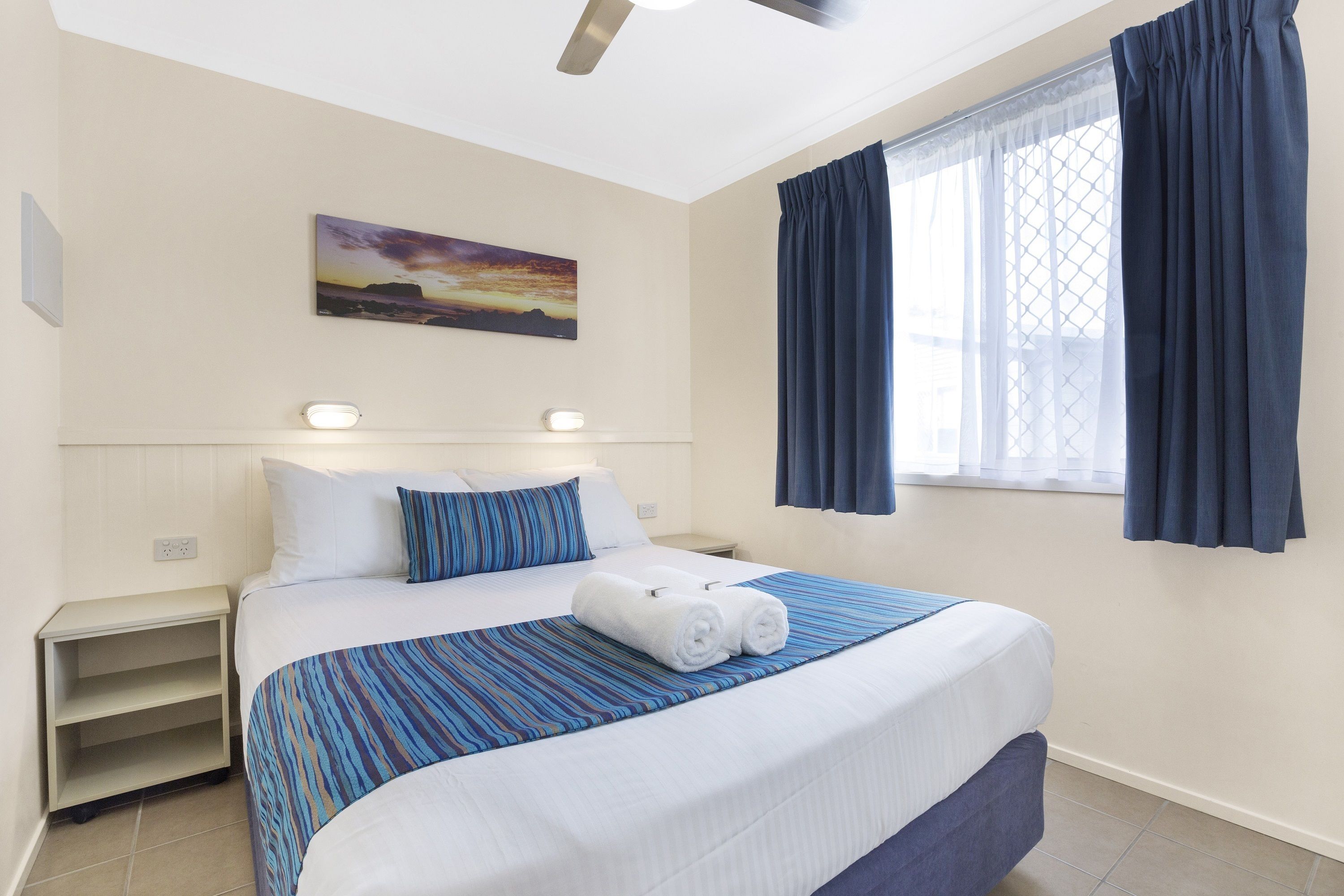 Quarto de hotel com cama, mesa de cabeceira, janela com cortinas, um quadro na parede e ventilador de teto.