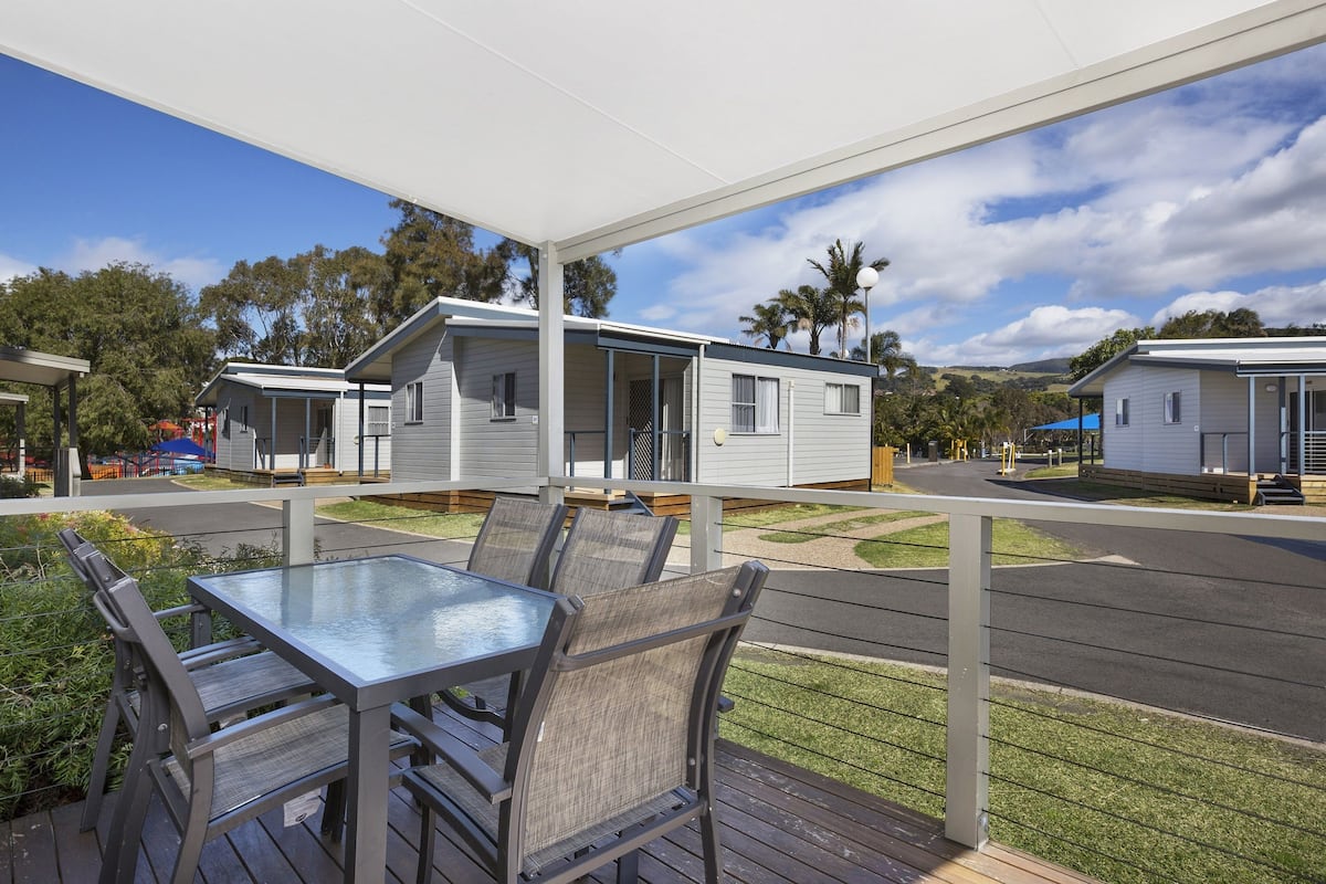 A covered outdoor area with a table and chairs, surrounded by mobile homes with a view of a road and trees.