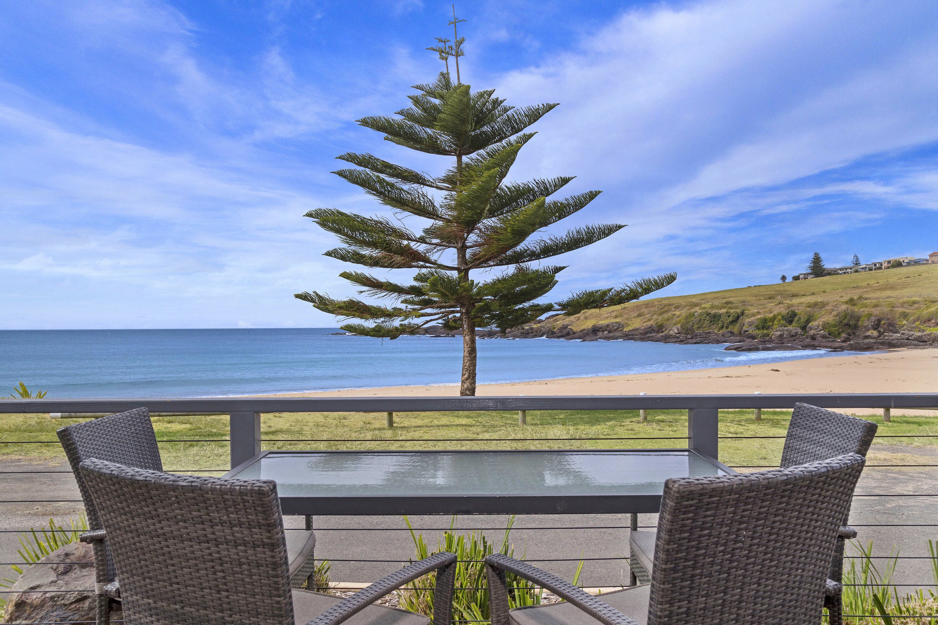 Une terrasse avec des chaises et une table en osier, donnant sur une plage et l’océan.